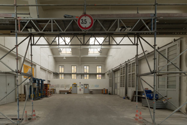 Großer Lagerraum mit ausgedehnten Gerüsten, Verkehrskegeln, Metallstangen, einem Schild, Maschinen, einer Tür, Fenstern, Wänden, Deckenleuchten und einem Dach.