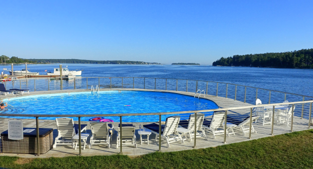 Großer Swimmingpool mit Liegestühlen und Whirlpool am Ufer eines Sees, umgeben von einem Geländer, mit Booten auf dem Wasser, Bäumen und einem klaren blauen Himmel im Hintergrund.