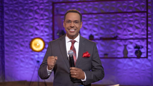 Stephen Colbert in Anzug, Krawatte und Brille auf einer Bühne mit Mikrofon, Wand, Ständer und Licht im Hintergrund sowie verschiedenen Gegenständen auf der Plattform.