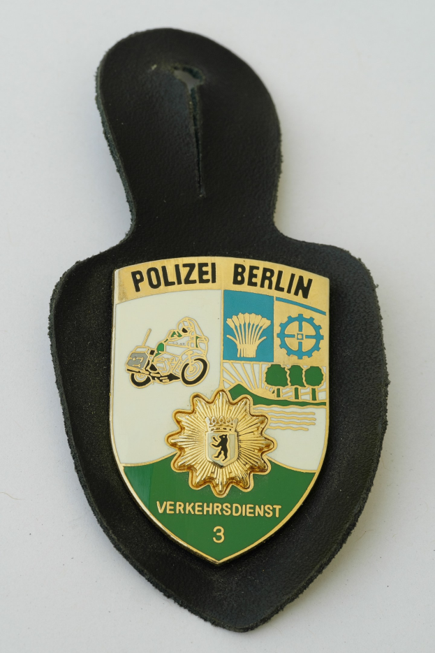 Ein schildförmiges Polizeisiegel mit 'Polizei Berlin' in schwarzer Fettschrift auf einem blauen Hintergrund mit einem weißen Stern und Rahmen, das auf einer Oberfläche liegt.