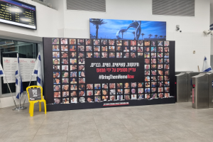 Große Wand mit mehreren Fotos, gelber Stuhl, israelische Regierungsflaggen auf Stangen, eine Tafel mit dem Text "Bring Them Home Now", ein montierter Bildschirm, Deckenleuchten und zusätzliche Objekte.