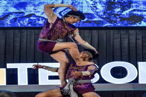 Zwei Akrobaten führen auf der Bühne eine Darbietung auf, wobei der Mann die Frau in lila Kleidern hochhebt, während das Publikum unten zuschaut.