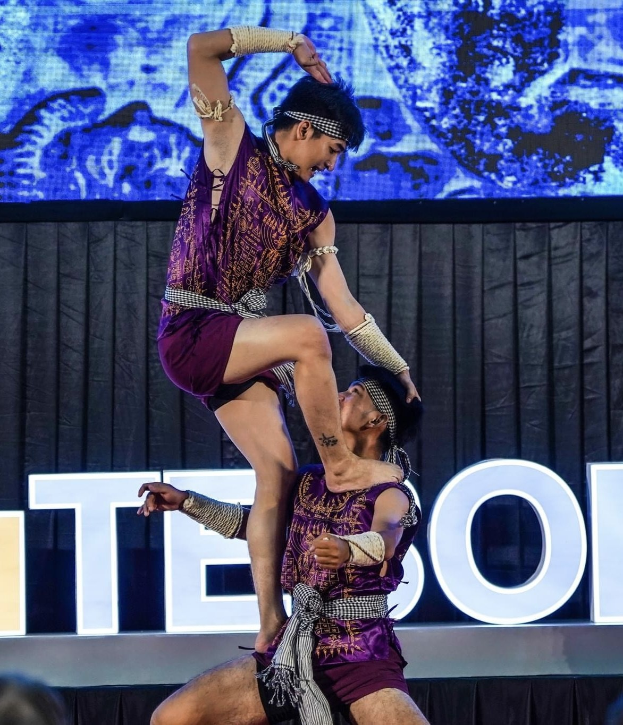 Zwei Akrobaten führen auf der Bühne eine Darbietung auf, wobei der Mann die Frau in lila Kleidern hochhebt, während das Publikum unten zuschaut.