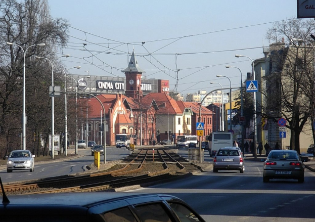 Eine belebte Stadtstraße mit Autos, Bussen, Straßenlaternen, Bahnschienen, Hinweistafeln, Laternenmasten, Bäumen, Gebäuden mit Fenstern und einem Uhrenturm unter einem klaren Himmel.