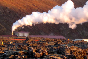 Ein Geothermiekraftwerk in Island mit Rauch, der aus seinem Schornstein aufsteigt, umgeben von Felsen im Vordergrund und H"ugeln im Hintergrund.