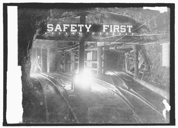 Schwaräz-weißes Foto eines Zuges, der aus einem Tunnel fährt, mit einer Person, die auf den Gleisen steht, im Vordergrund, Hintergrund zeigt Strommasten, Lichter und ein "Sicherheit zuerst"-Schild.