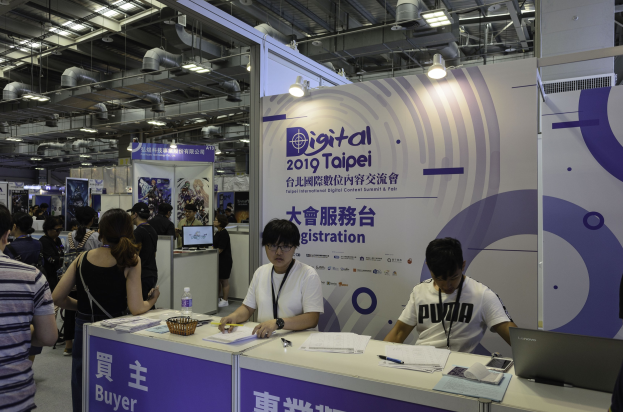 Messebesucher an einem Messestand für die Digital 2019 Taipei International Digital Content Summit & Fair, mit zwei sitzenden Personen vor einem Tisch mit Büromaterialien und -artikeln.