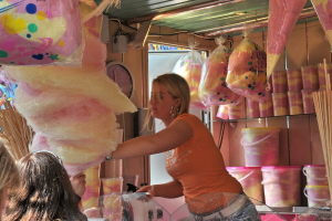 Eine Frau steht vor Glasregalen mit Päckchen und Gläsern voller Lebensmittel, mit einer Uhr an der Wand darüber und einer Leuchte über der Uhr und Päckchen mit Süßigkeiten links daneben, während Köpfe von Menschen in der unteren linken Ecke sichtbar sind.