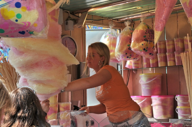Eine Frau steht vor Glasregalen mit Päckchen und Gläsern voller Lebensmittel, mit einer Uhr an der Wand darüber und einer Leuchte über der Uhr und Päckchen mit Süßigkeiten links daneben, während Köpfe von Menschen in der unteren linken Ecke sichtbar sind.
