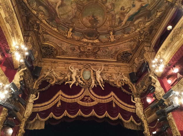 Theaterinnenraum mit einer roten Gardine, Kronleuchtern, Skulpturen, Lichtern, einer Deckenmalerei und einer Bühne mit ein paar Menschen unten.