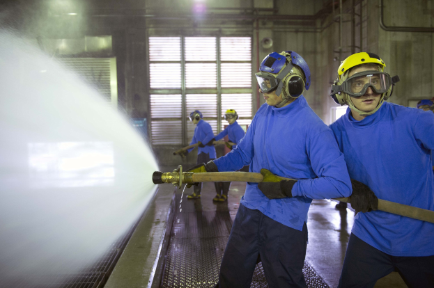 Gruppe von Männern in blauen Hemden und gelben Helmen, die an Maschinen arbeiten, wobei einer Wasser auf den Boden sprüht, in einer Fabrik mit sichtbaren Rohren, Fenstern und Lampen.