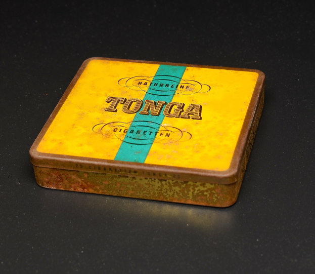 Ein gelb-blauer Blechbehälter mit Tonga Zigaretten auf einem schwarzen Tisch, mit Text, der die Marke und Sorte des Tabaks angibt.