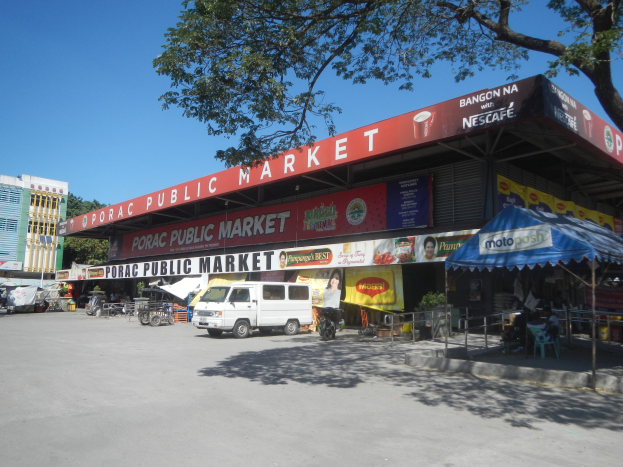 Öffentlicher Markt mit einem weißen Van vorne, umgeben von Gebäuden, Motorfahrzeugen, Menschen, die unter einem Zelt sitzen, Geländern, Bäumen und einem klaren blauen Himmel.