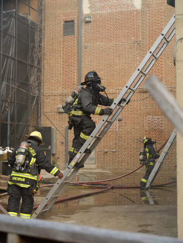 Feuerwehrleute in Helmen und Ausrüstung klettern an einer Leiter vor einem Backsteingebäude mit Rohren auf dem Boden und einer Metallstange am unteren Ende hoch, wobei im Hintergrund ein weiteres Gebäude mit Fenstern und einem Netz zu sehen ist.