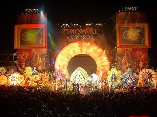 Eine große Menschenmenge bei Nacht vor einer Bühne mit farbigen Lichtern, auf der ein Bildschirm mit der Aufschrift "Carnaval de Vinaros" zu sehen ist.