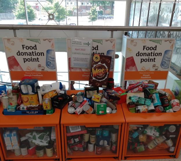 Orange Schübe mit Lebensmitteln wie Flaschen, Dosen und Kisten in einer Spendenstelle, mit Informationsschildern und einer Aussicht auf Fahrzeuge, Bäume, Gebäude und Himmel durch Glasfenster.