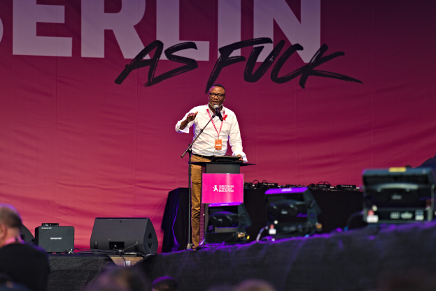 Ein Mann in weißem Hemd und braunen Hosen steht auf einer Bühne und hält ein Mikrofon in der Hand, hinter ihm eine pinkfarbene Banner mit der Aufschrift 'Berlin as Fuck' und Lautsprecher auf der Bühne.