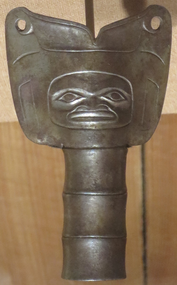 Nahaufnahme eines Messers aus Metall mit einem geschnitzten Gesicht am Griff, das auf einem Holztisch gegen eine Holzwand gestellt ist.