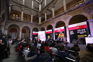 Eine Gruppe von Menschen sitzt auf einer Pressekonferenz für das CDMX 2017-Event, mit einem Podium und einem Mikrofon in der Mitte, umgeben von einer Menge mit Kameras, Schildern, Säulen, Geländern, Fenstern und einer Wand im Hintergrund.