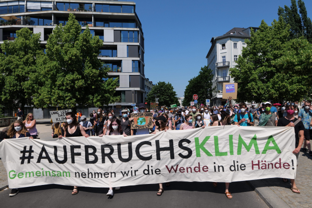 Eine Gruppe von Menschen mit Masken, die ein Transparent mit der Aufschrift 'Aufbruchsklima' vor einem Gebäude mit Bäumen und einem klaren Himmel halten.