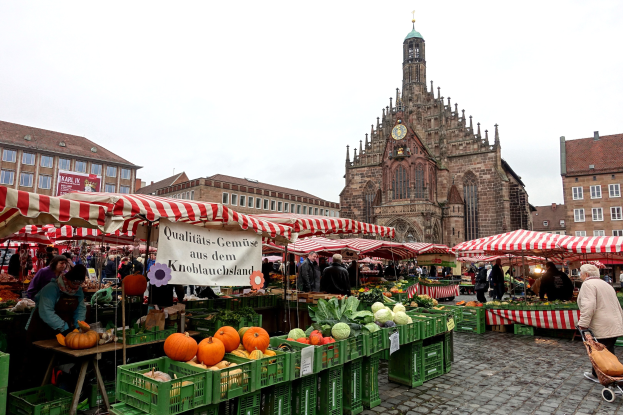 Ein belebter Markt in Nürnberg, Deutschland, mit Ständen voller Obst und Gemüse, Menschen, die einkaufen, und aufgestellten Zelten, mit Gebäuden und einem Kirchturm im Hintergrund unter einem sichtbaren Himmel.