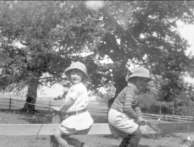 Zwei junge Jungs in Mützen mit Baseballschlägern in einem Park mit Bäumen und einem Zaun im Hintergrund, in Schwarz-Weiß.