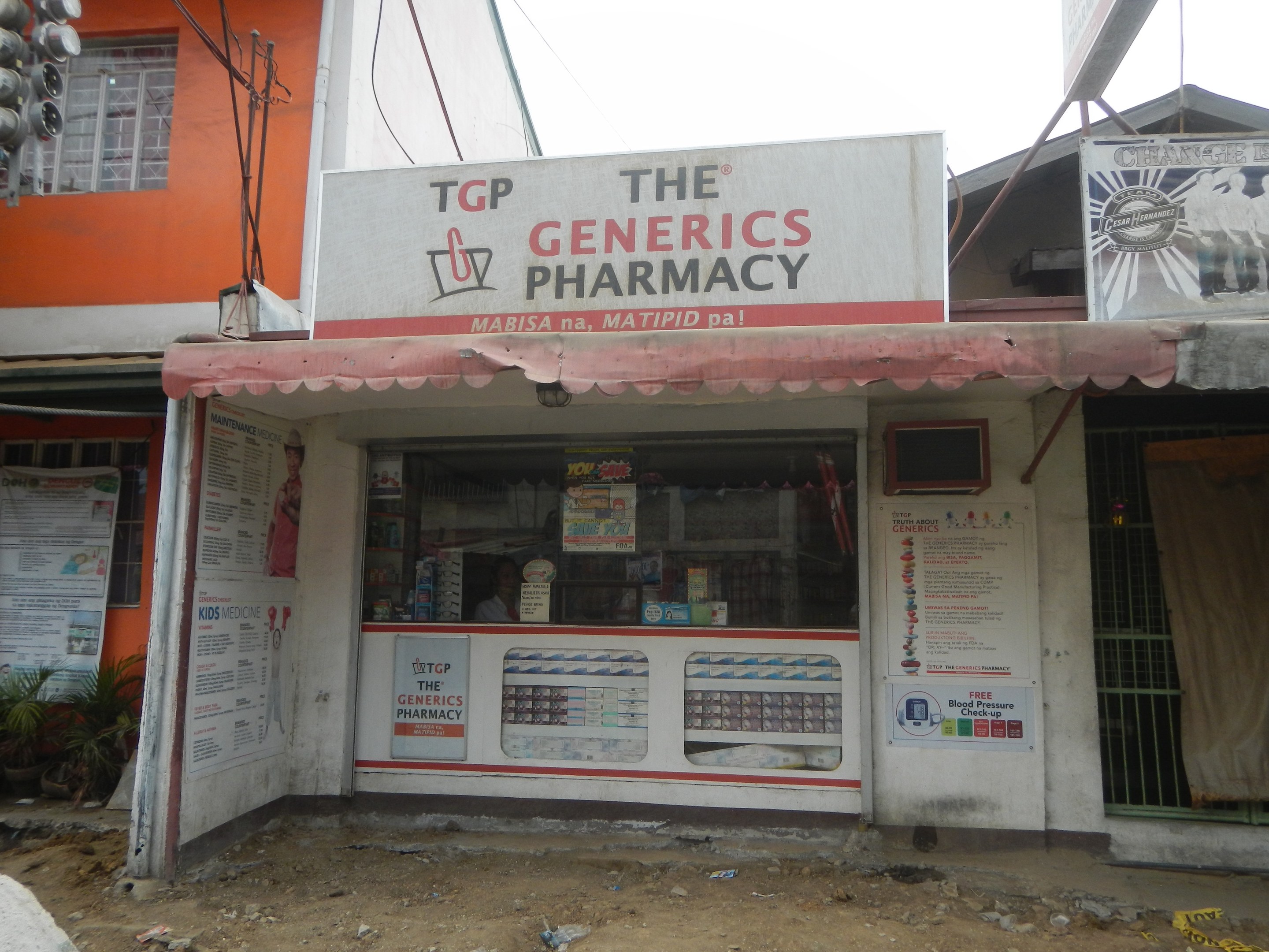 Außensicht der The Generics Pharmacy Filiale mit umliegenden städtischen Elementen unter einem klaren blauen Himmel.
