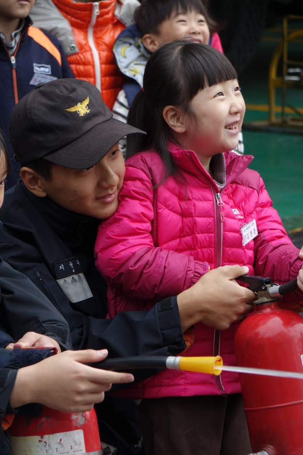 Eine Gruppe von Kindern steht um einen roten Hydranten in einer Schulumgebung, wobei eines der Kinder ein Rohr hält.