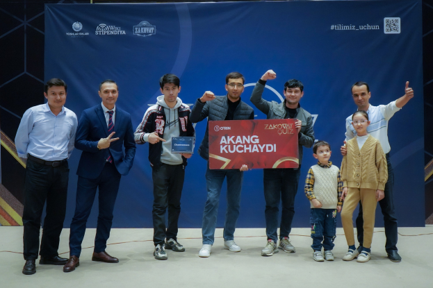 Gruppe von Menschen auf einer Bühne mit einer Tafel, auf der "Akang Kuchaydi - Zakovat Quiz" steht, im Hintergrund ein Banner, Strichcode und Beleuchtung.