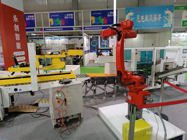 Eine Gruppe von Robotern sitzt an einem Tisch in einer Fabrikumgebung umgeben von Maschinen, Kabeln, Texttafeln und Lichtern, mit Menschen im Hintergrund.