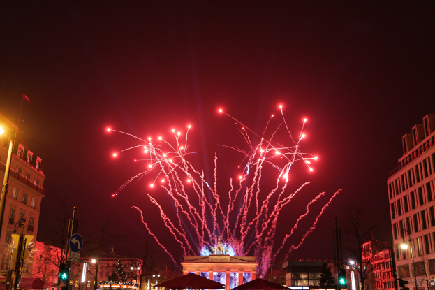 Eine belebte Straße in Berlin an Silvester, geschmückt mit Gebäuden, Bäumen, Laternenmästen, Verkehrszeichen, Schildern, Zelten und Menschen, vor einer Kulisse von Feuerwerk, das den Himmel erleuchtet.