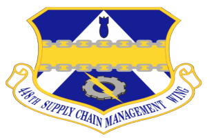 Ein Schild mit einem blauen Hintergrund, ein weißer Stern in der Mitte und ein weißes Banner mit den Worten "Supply Chain Management" in blau geschrieben, umgeben von einem weißen Rand, mit dem Text "48th Supply Chain Management Wing" in fetter, schwarzer Schrift.