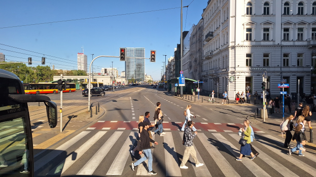Menschen, die eine Straße an einer Ampel überqueren, mit einem Bus auf der linken Seite, Fahrzeugen auf der Straße, Verkehrssignalen, Laternenmasten, Gebäuden, Bäumen und einem klaren blauen Himmel.