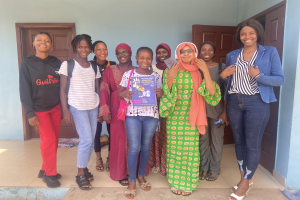 Eine Gruppe von Frauen, die vor einem Schulgebäude in Nigeria lächeln und Bücher halten, mit einem Tuch auf dem Boden und Türen im Hintergrund.