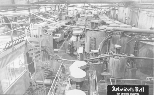Schwarzes und weißes Foto eines großen Fabrikinneren mit einer Person, die zwischen Industriegeräten, Rohren und baulichen Elementen steht, mit dem Text "Arbeidets Rett, die größte Fabrik der Welt" unten.