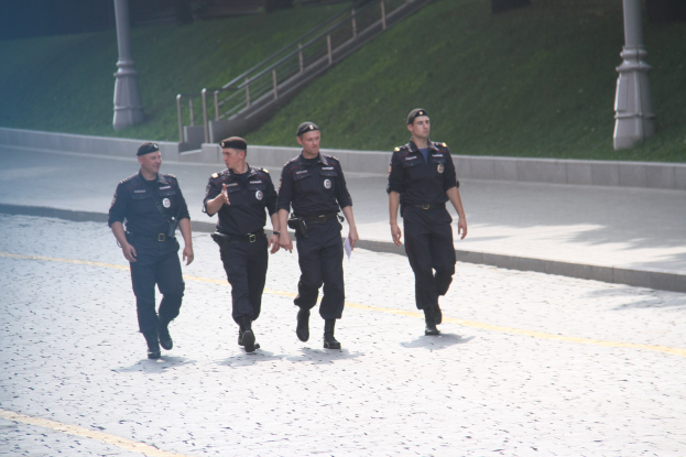 Drei Polizisten in Uniform und Mützen, die eine Straße mit Pfosten, Geländern, Treppen und Gras im Hintergrund entlanggehen.