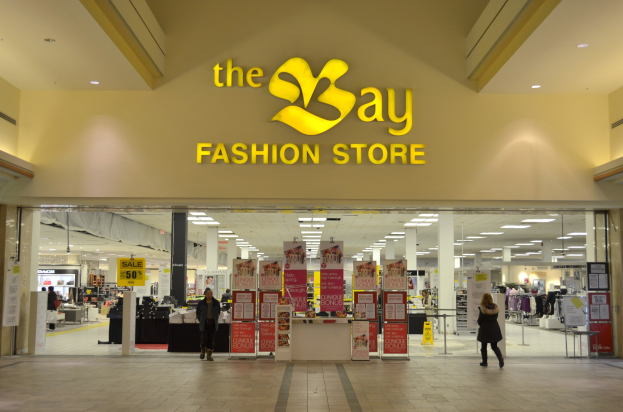 Ein Modegeschäft namens "bay" in einem Einkaufszentrum mit Menschen, die gehen, Texttafeln an den Wänden, Säulen, Deckenleuchten und Hintergrundobjekte.