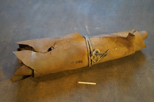 Ein brauner Papiertütenbeutel in Form einer Granate mit einem Griff oben drauf, mit einem Seil zugebunden, und eine Zigarette daneben auf einem Tisch.