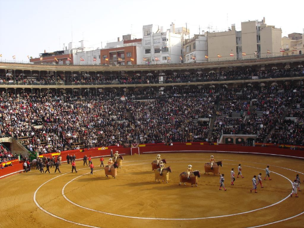 Menschen zu Fuss auf dem Boden, einige reiten Pferde, und eine Menge Menschen sitzen im Stadion mit Fahnen, Gebäuden und Himmel im Hintergrund.