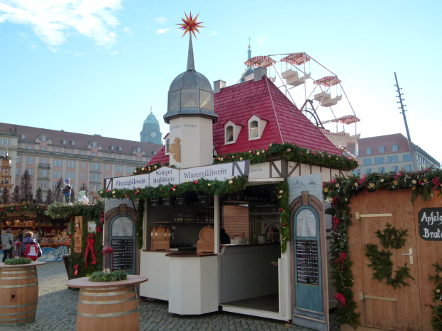 Kleines Gebäude mit rotem Dach und Riesenrad davor, umgeben von Menschen und Festobjekten, mit Gebäuden, Bäumen und bewölktem Himmel im Hintergrund.