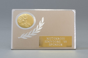 Eine goldene und silberne Visitenkarte mit Lorbeerkranz und Text, mittig auf einem einfarbigen Hintergrund.