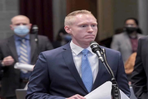 Ein Mann im Anzug und Krawatte spricht an einem Rednerpult mit Papieren, während andere hinter ihm stehen.