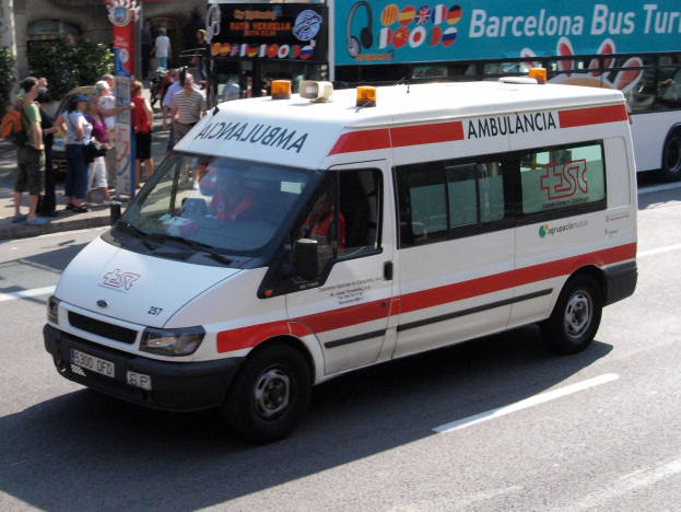 Rettungswagen fährt an einem Bus mit Menschen auf dem Gehweg vorbei, Gebäude und Bäume im Hintergrund unter einem klaren blauen Himmel.