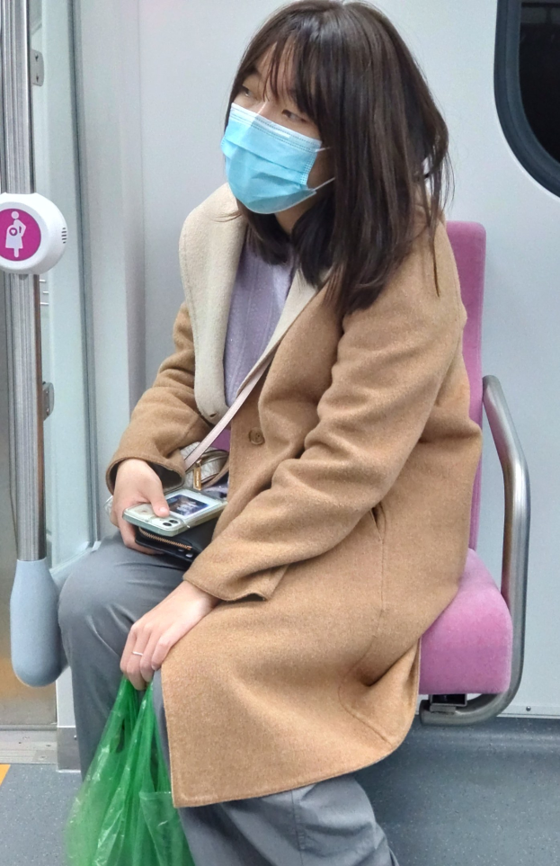 Eine Frau mit Gesichtsmaske sitzt in einem Zug, hält ein Mobiltelefon und eine Tasche und hat eine Tür links von sich.