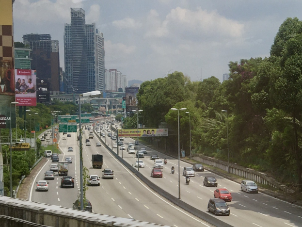 Eine verkehrsreiche Autobahn mit hohen Gebäuden, Straßenlaternen, Leuchtreklamen, Bäumen und einem bewölkten Himmel.