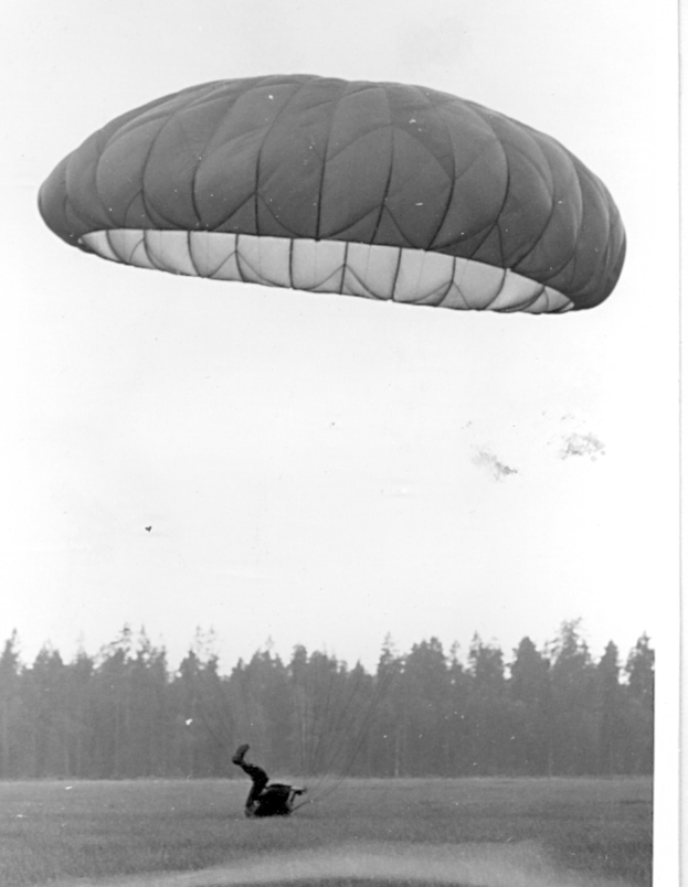 Schwarzes und weißes Foto einer Person beim Paragleiten mit einem Fallschirm, umgeben von Bäumen und einem klaren Himmel, mit Text unten.