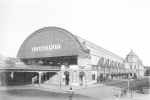 Ein Schwarz-Weiß-Foto von Alexanderplatz in Berlin, Deutschland, das ein großes Gebäude mit Säulen, Bögen und einem Namensschild, Laternen und Menschen auf der Straße zeigt, mit Bäumen und einem klaren Himmel im Hintergrund.