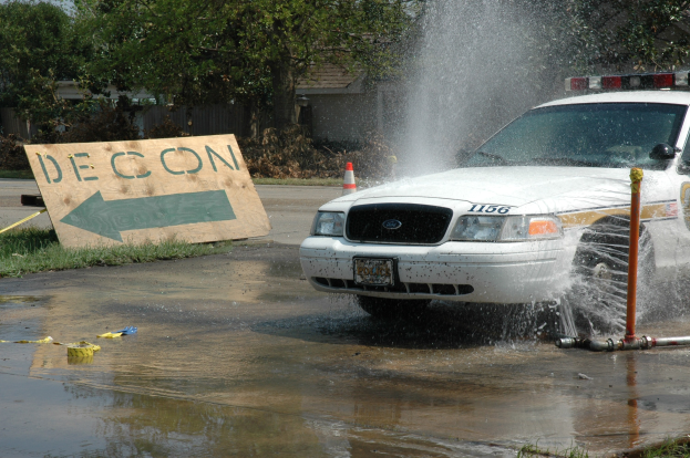 Polizeiauto fährt durch eine Pfütze mit verstreuten Gegenständen einschließlich einer Platte, einer Verkehrsinsel, Bäumen, Gebäuden und einem klaren blauen Himmel im Hintergrund.