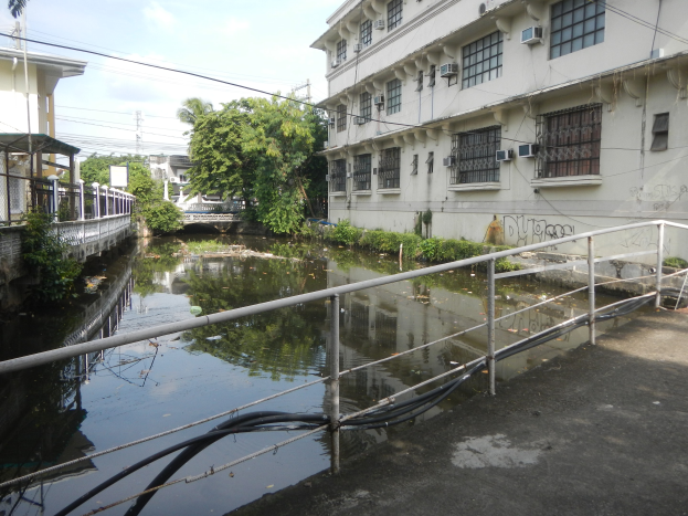 Ein kleiner Kanal verläuft durch eine Stadtstraße, flankiert von hohen Gebäuden, mit Wasser, Geländern, Rohren, Pflanzen, Bäumen, Strommasten mit Drähten und einem bewölkten Himmel im Hintergrund.