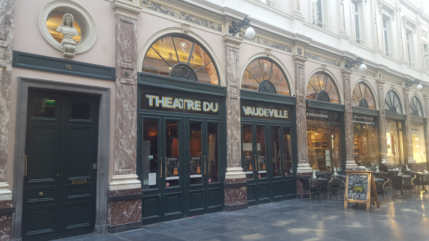 Theater du Vaudeville-Gebäude in Paris, mit Türen, Fenstern, beleuchteter Beschilderung, Tischen, Stühlen und einer Wandstatue.